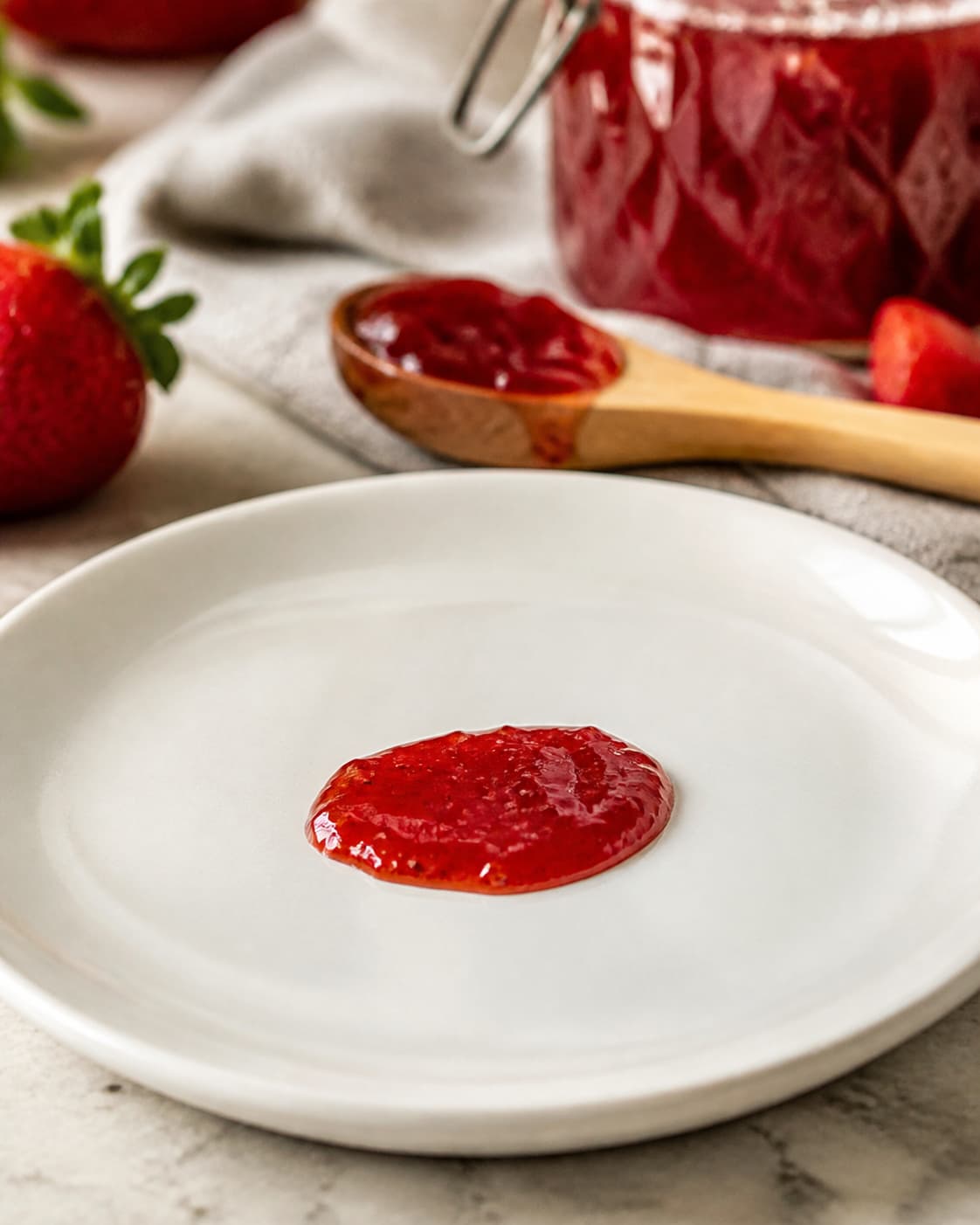 Une petite goutte de confiture de fraise rouge brillante posée sur une assiette en céramique blanche froide, légèrement ridée, avec une cuillère en bois floutée en arrière-plan et un éclairage chaleureux de cuisine.