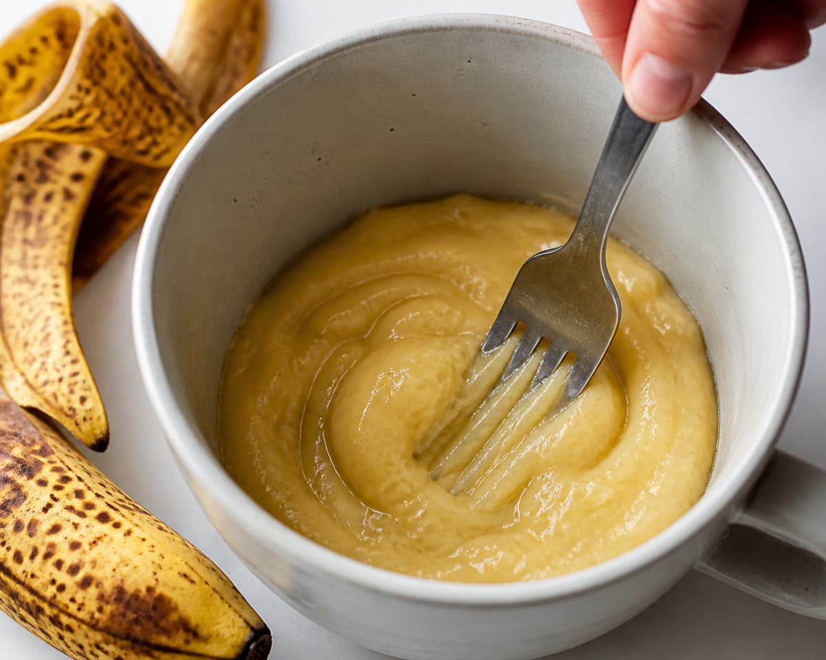 Fourchette écrasant une banane très mûre tachetée au fond d’un mug, formant une purée lisse.