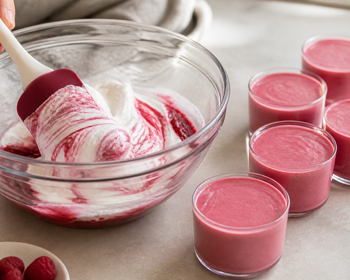 Spatule souple incorporant délicatement une crème montée dans une purée de framboises, créant un mélange marbré, avec des verrines remplies de mousse lisse alignées à côté.