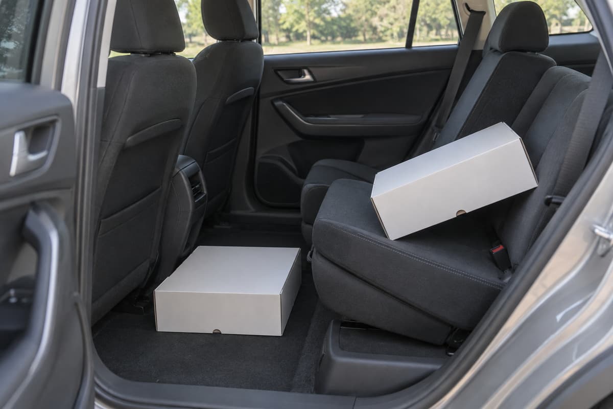 Boîte à gâteau posée à plat sur le plancher arrière d’une voiture, à côté d’un siège incliné sur lequel une boîte similaire est en équilibre précaire, avec un espace dégagé au-dessus.