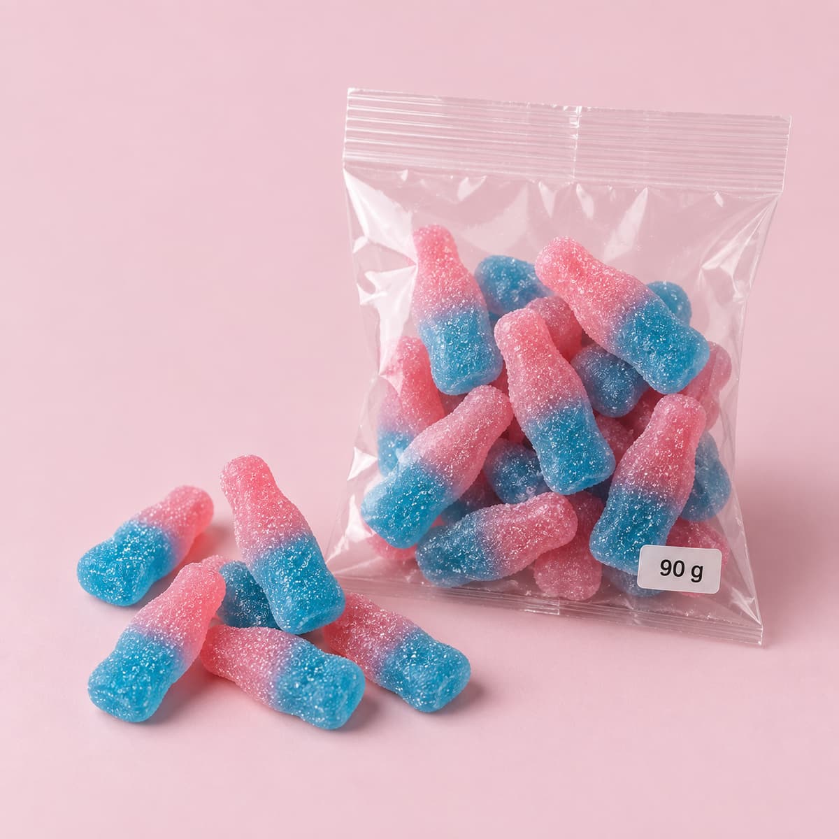 Bonbons en forme de bouteille bicolores rose et bleu posés à côté d’un sachet transparent indiquant 90 g sur fond clair.
