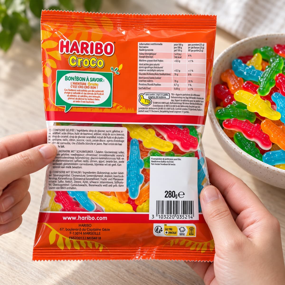 Deux mains tiennent le dos d’un sachet de bonbons près d’un bol contenant les mêmes confiseries, un doigt pointant la liste des ingrédients sans la masquer.