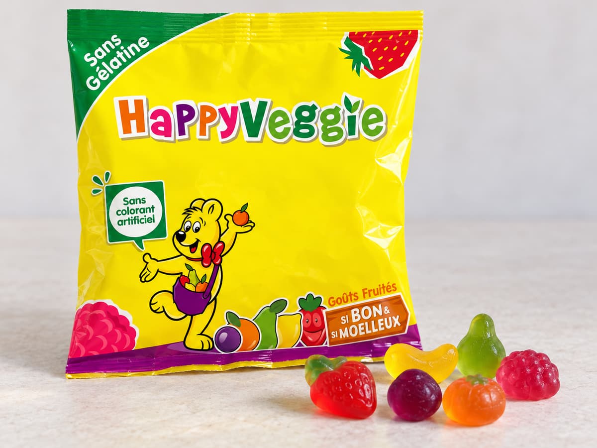 Sachet de bonbons HARIBO Happy Veggie posé sur une surface claire avec plusieurs bonbons colorés de formes variées devant lui.